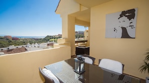 A balcony with a table and chairs, a view of the landscape, and a painting on the wall. - Duquesa Village 8.25.21 (Manilva)