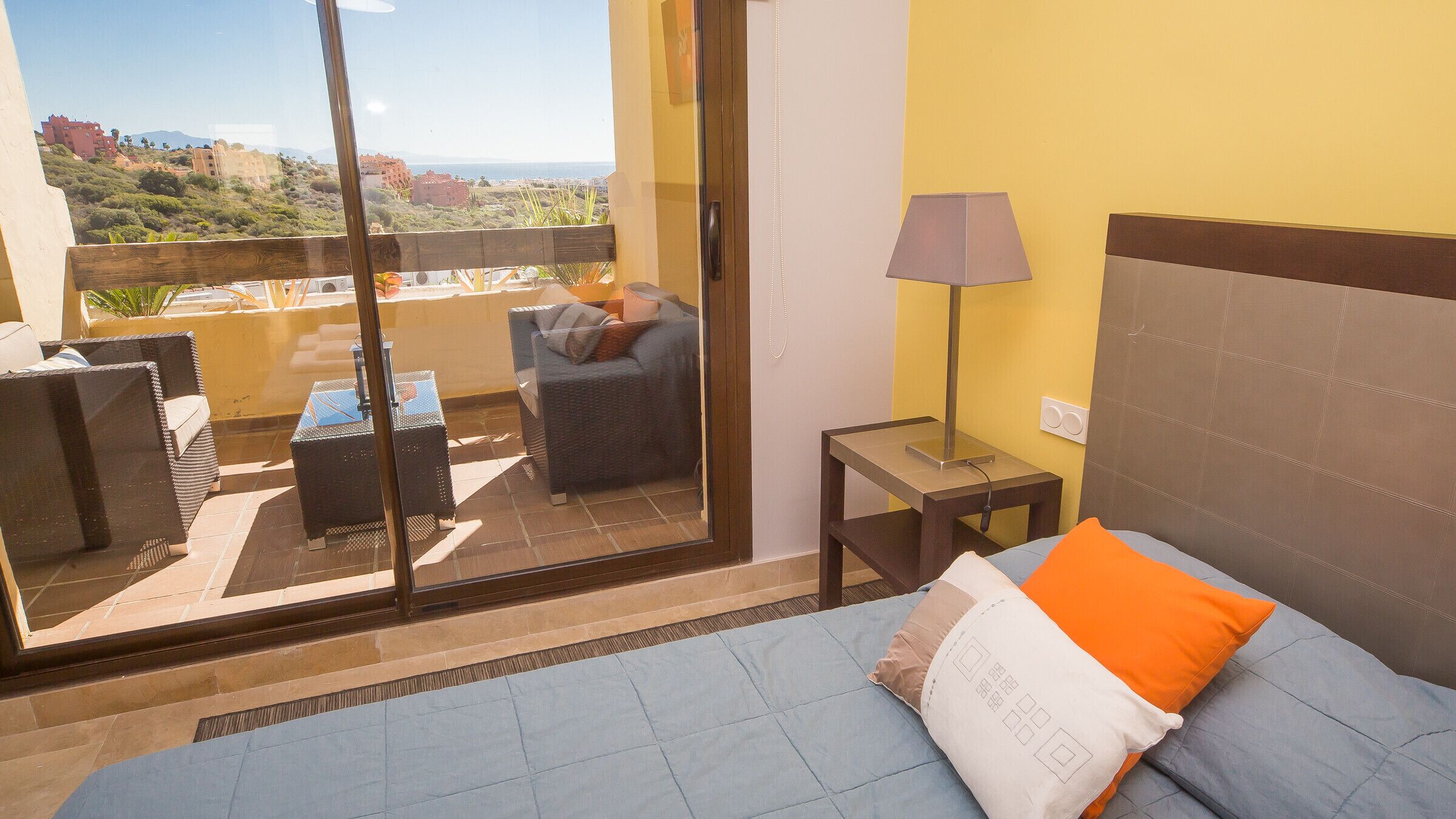 Uma varanda com sofá e mesa, uma cama com travesseiro laranja e um abajur.