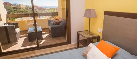 Uma varanda com sofá e mesa, uma cama com travesseiro laranja e um abajur.
