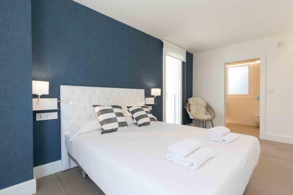 A hotel room with a large bed, two bedside lamps, a chair, and a view of the bathroom. - Brunet III | The Rentals Collection (San Sebastián)