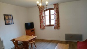 A room with a wooden dining table and chairs, a television on a wooden cabinet, a window with patterned curtains, and a radiator. - 2-Room Apartment for 4 in Vignec (Vignec)