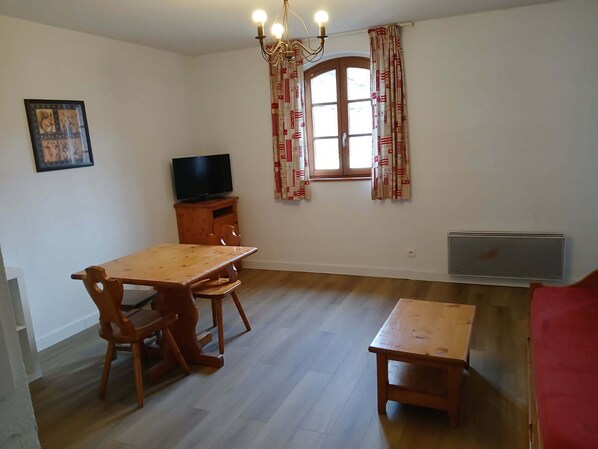 A room with a wooden dining table and chairs, a television on a wooden cabinet, a window with patterned curtains, and a radiator. - 2-Room Apartment for 4 in Vignec (Vignec)