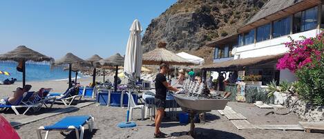 A beach with sun loungers, umbrellas, and a building with a thatched roof.