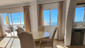 A dining area with a table and chairs, large windows with curtains, a view of the sea, and a kitchen area with a dishwasher. - Spacious and bright penthouse with panorama seaview (Nerja)