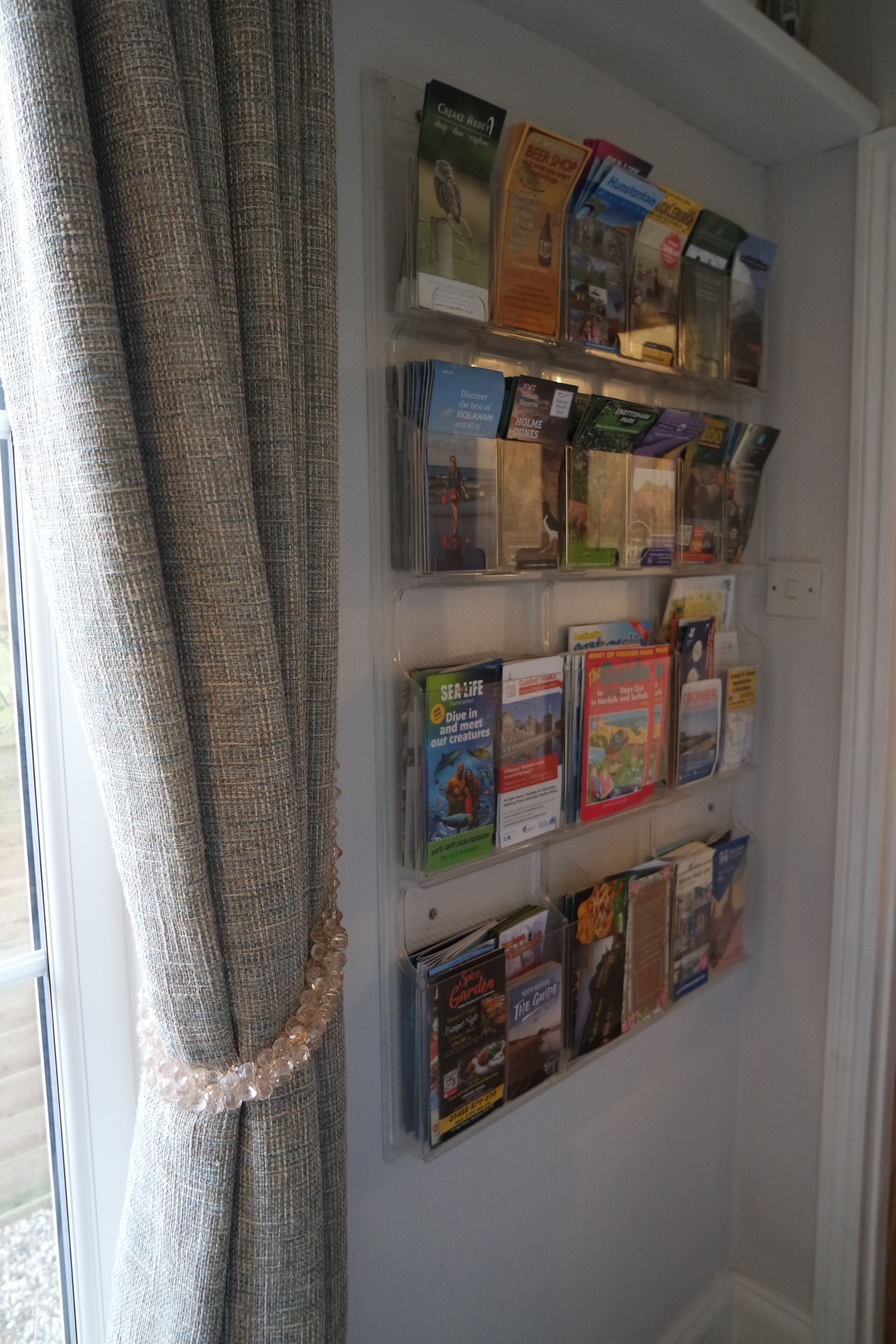 Un mur orné d’une collection de livres et de magazines, une fenêtre avec un rideau et une étagère fixée au mur, sur laquelle sont disposés divers objets.