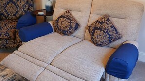 Un salon avec un canapé beige, des coussins bleus, un fauteuil à motifs, une table d’appoint avec une lampe et un panneau d’accueil encadré.