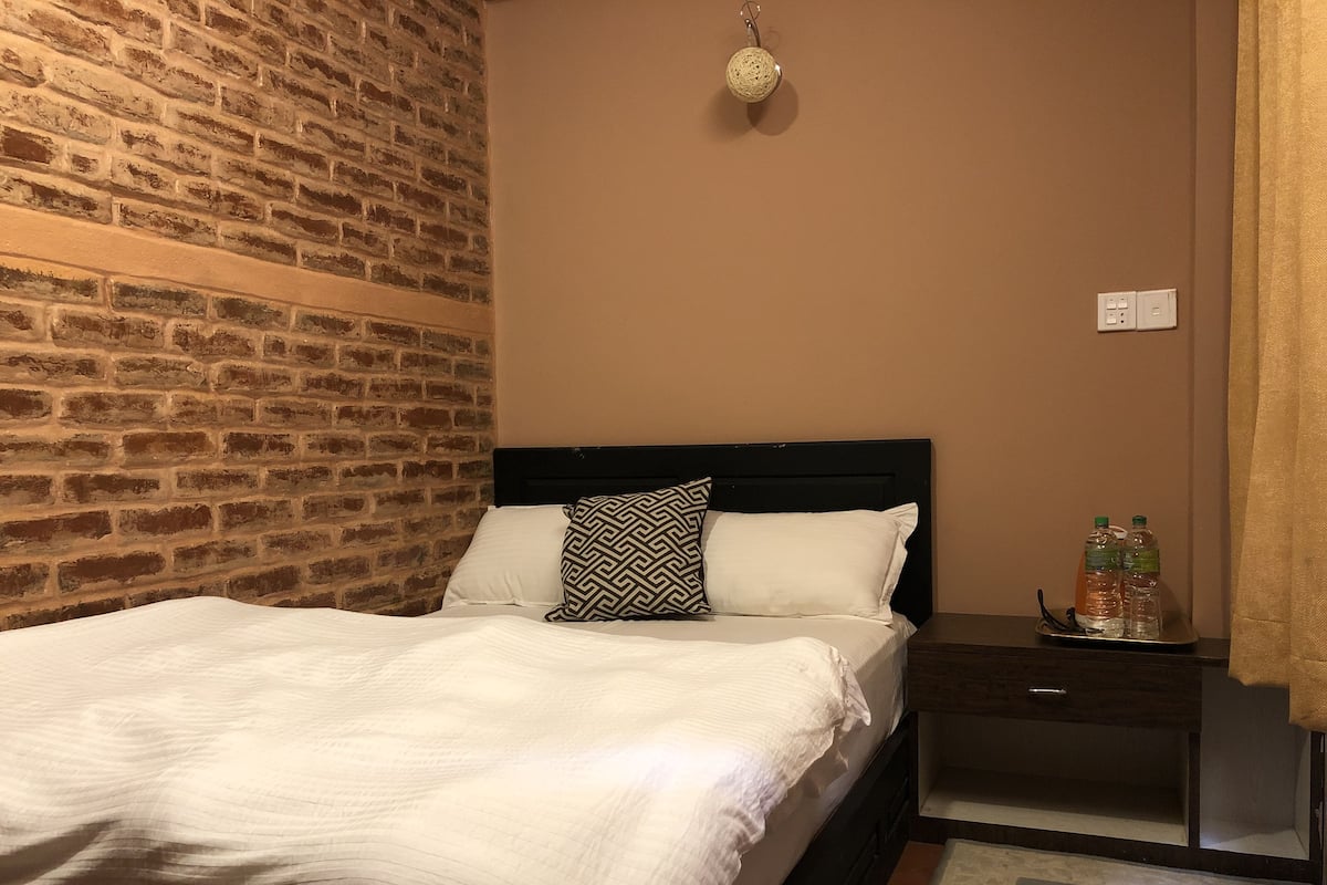 Una cama bien hecha con un cojín estampado, una mesita de noche con frascos y una pared de ladrillo.