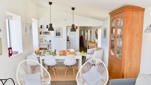 A modern living room with a grey sofa, two white wicker chairs, a wooden cabinet, and a dining area with a table and chairs.