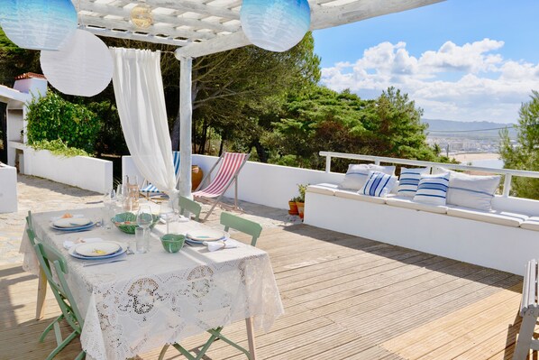 An outdoor dining area with a white table set for four, surrounded by a wooden deck and overlooking a scenic landscape.