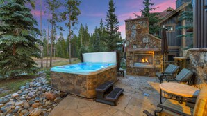 A hot tub with stone steps, surrounded by outdoor seating and a stone fireplace.