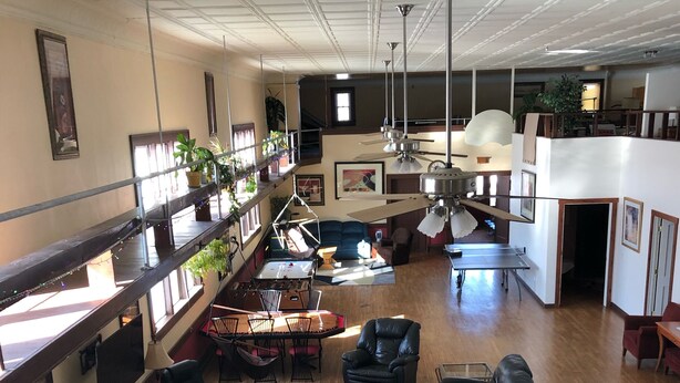 Amplia sala de estar con techo alto, múltiples opciones de asientos que incluyen sofás y sillones, una mesa de centro y una mesa de comedor. Hay plantas en macetas y obras de arte en las paredes, además de un balcón con barandillas.