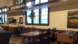 Una habitación amplia con una mesa de madera, sillas, una mesa de futbolín y un gran ventanal con plantas.