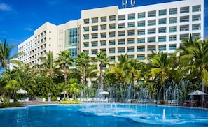 Een hotel met een groot zwembad, palmbomen en een moderne gevel.