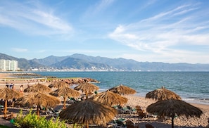 Een strand met rieten parasols, een zandstrand en bergen op de achtergrond.