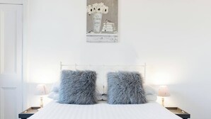 Ett sovrum med en vit säng, två fluffiga kuddar, ett vitt nattduksbord med en lampa och en tavla med blommor och böcker som hänger på väggen.