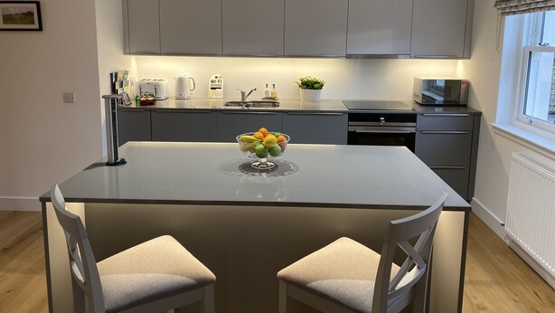 A modern kitchen with grey cabinets, a central island with a bowl of fruit, a microwave, and a sink. - Luxury flat (St Andrews)