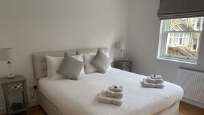A neatly made bed with pillows, a bedside table with a lamp, and a window with a view of a house. - Luxury flat (St Andrews)
