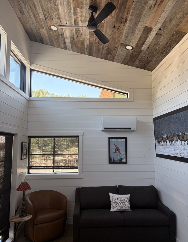 A room with a ceiling fan, a window with a view, a couch, and a wall-mounted air conditioner. - Newly Built Shady Creek Tiny Home @ Tiny Timbers Resort! (South Fork)