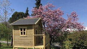Una casita de juegos de madera con escalera y tobogán, rodeada de árboles florecientes y césped.