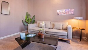 Ein modernes Wohnzimmer mit grauem Sofa, einem Glastisch, einer großen Grünpflanze und einer Wandlampe.