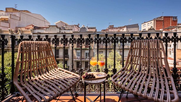 Ein Balkon mit zwei Korbstühlen, einem kleinen Tisch mit einer Schale Kirschen und zwei Gläsern Orangensaft.