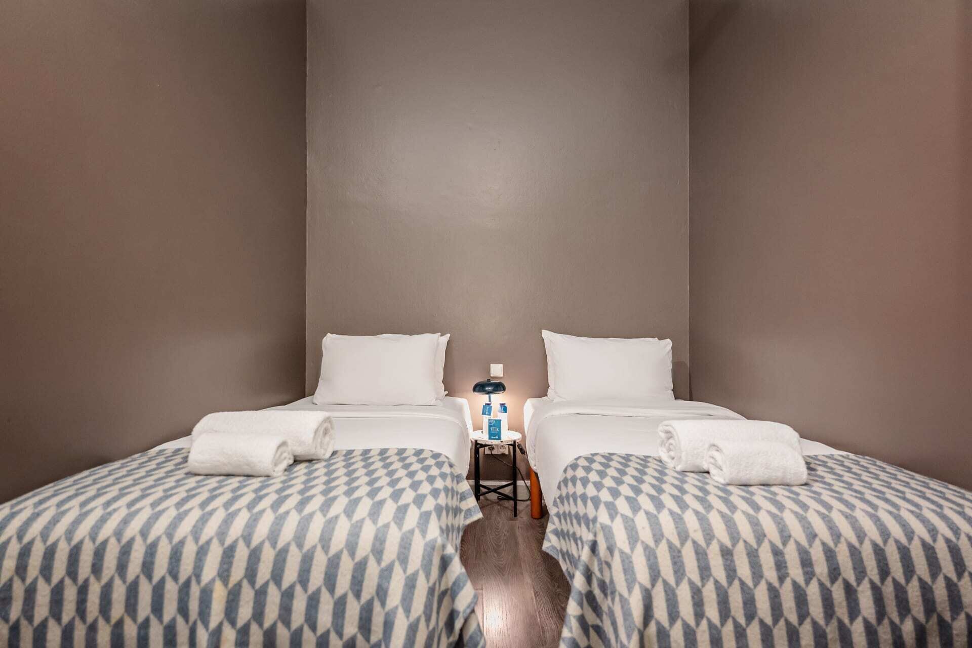 A hotel room with two beds, each with a patterned bedspread, a small table with toiletries, and a nightstand with a lamp.