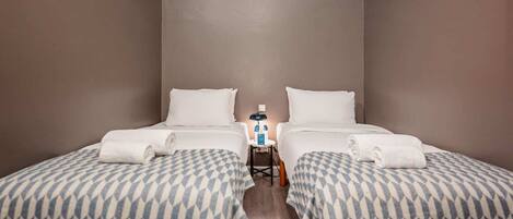 A hotel room with two beds, each with a patterned bedspread, a small table with toiletries, and a nightstand with a lamp.