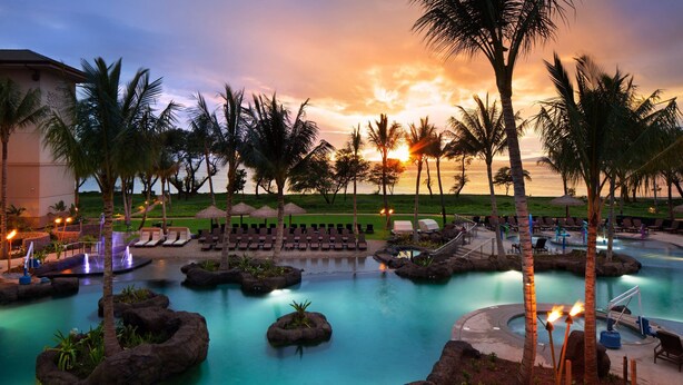 Un espace piscine aménagé dans un complexe hôtelier, avec des chaises longues, des palmiers et une vue sur le coucher de soleil.