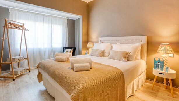 Une chambre d’hôtel avec un grand lit, une table de chevet avec une lampe, une échelle en bois avec un panier, et une fenêtre avec des rideaux légers.