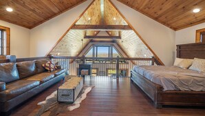 A spacious loft bedroom with a sloped ceiling, a wooden bed, a leather sofa, and a view of the outdoors. - Peregrine's Perch- amazing views, hot tub, outdoor fireplace, Eagles Nest amenities (Banner Elk)
