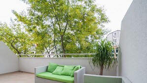 A balcony with a wicker sofa and a potted plant, overlooking a tree-lined area. - Comfortable apartment located in the quier neighborhood in a new building . (Budva)