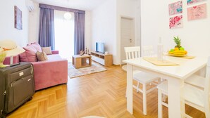 A modern living room with a pink sofa, a wooden coffee table, and a white dining table. There is a TV on a stand, a suitcase, and a fruit bowl on the dining table. - Comfortable apartment located in the quier neighborhood in a new building . (Budva)