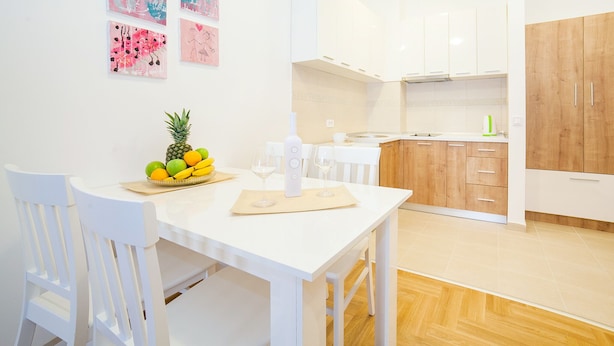A modern kitchen with white cabinets, a wooden cabinet, and a dining area with a white table and chairs. - Comfortable apartment located in the quier neighborhood in a new building . (Budva)