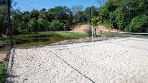 Uma quadra de vôlei de areia com rede, cercada por árvores e um pequeno lago.