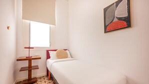 Ein kleines, ordentlich eingerichtetes Schlafzimmer mit einem Einzelbett, einem Nachttisch, einer Stehlampe und einem Fenster mit Jalousie.