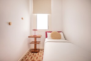 Een kleine, verzorgde slaapkamer met een bed, een nachtkastje, een lamp en een raam met een jaloezie.