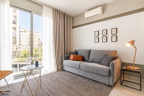 A modern living room with a grey sofa, a small round table, a floor lamp, and a view of buildings through large windows.