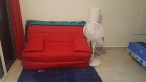 A red sofa with a patterned cover, a standing fan, and a blue mat on a tiled floor. - Studio any comfort. (Terre de haut les saintes)