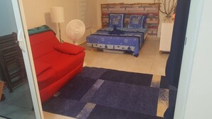 A bedroom with a red sofa, a bed with a blue bedspread, a standing fan, and a brick-patterned headboard. - Studio any comfort. (Terre de haut les saintes)