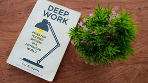 A book titled 'DEEP WORK' by Cal Newport on a wooden table next to a small potted plant with pink flowers.
