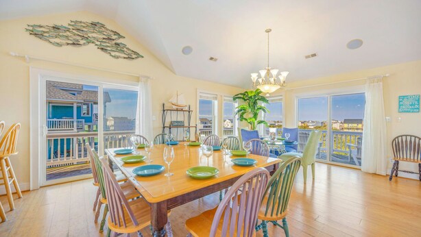 A spacious dining area with a large wooden table, colorful chairs, and a view of the sea through large windows. - Oceanview Dog Friendly with Private Pool, Hot Tub, Elevator, Game Room, Theater (Rodanthe)