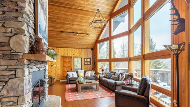 A spacious living room with a stone fireplace, wooden ceiling, large windows, and a cozy seating area. - Mountaineer Manor- views, hot tub, 3 acres fenced in yard, pool table (Sugar Grove)