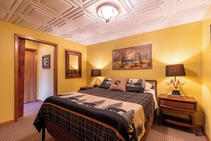 Un dormitorio con una cama, dos lámparas de noche, una mesita de noche, un cuadro en la pared y una puerta que da a otra habitación.