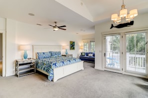 A bedroom with a bed, a ceiling fan, a chandelier, a large window with blinds, and a door leading to a balcony.