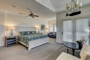 A bedroom with a bed, bedside tables, a ceiling fan, a chandelier, a large window with blinds, and a fish decoration on the wall.