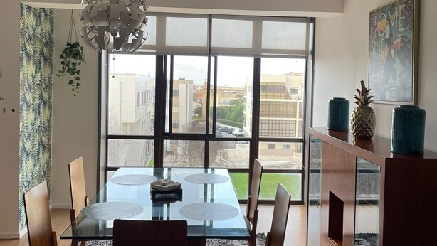 Una sala moderna con una mesa de comedor de vidrio, sillas de madera, un sofá y un amplio ventanal con vistas a la ciudad.