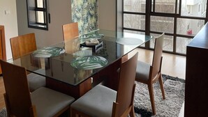 Un comedor moderno con una mesa de vidrio, sillas de madera, una araña y un ventanal con vista a la ciudad.