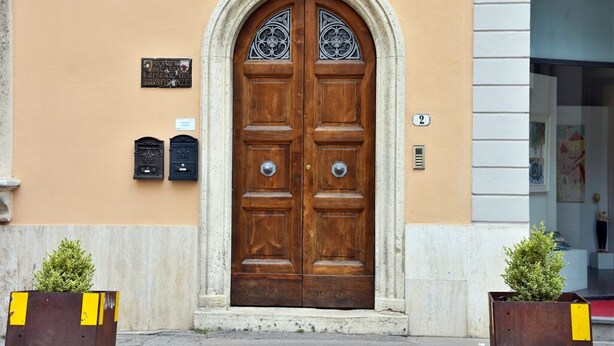 A double wooden door with a stone archway, a plaque, and two mailboxes on the wall. - - Piazza Arnolfo 1932- (Colle di Val d'Elsa)