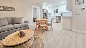 A modern living room with a sofa, a round coffee table, and a dining area with a table and chairs. There is a kitchen in the background with white cabinets and stainless steel appliances. -  Beachy Keen Cottage B!  Just Steps from Indian Rocks Beach! (Indian Rocks Beach)