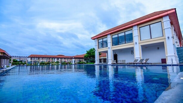 A swimming pool with lounge chairs and a building with large windows and a red roof. - Spoorti Resort and Club (Bijapur)